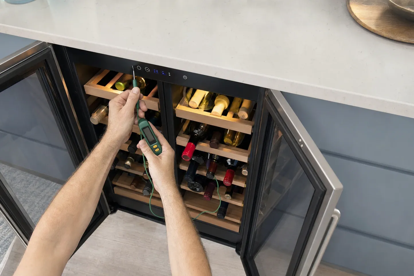 Overhead view of hands lowering a probe thermometer into an open undercounter wine cooler, bottles resting on wooden racks